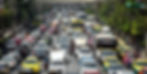 Heavy traffic on an urban street with cars, buses, and motorcycles in various colors. Trees line the road. Buses and taxis dominate.