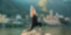 A woman doing yoga on a rock by a river, with colorful buildings and lush hills in the background, creating a serene atmosphere.