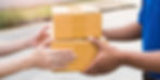 Hands exchanging two brown cardboard boxes outdoors. One person wears a blue shirt. Bright, natural lighting suggests a daytime setting.