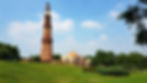 Qutub Minar - Tall sandstone tower with intricate design in a green park under a blue sky. People walk around the lush grass and trees. Historic architecture.